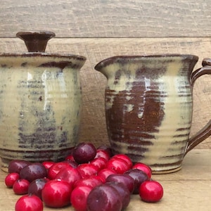May include: A set of two handmade ceramic kitchenware pieces in a brown and cream glaze. The set includes a pitcher and a lidded jar.