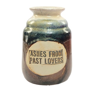 Könnte beinhalten: Eine braune und weiße Keramikvase mit rundem Körper und einer schmalen Öffnung. Die Vase hat ein weißes ovales Etikett mit dem Text "Ashes from Past Lovers" in Schwarz.
