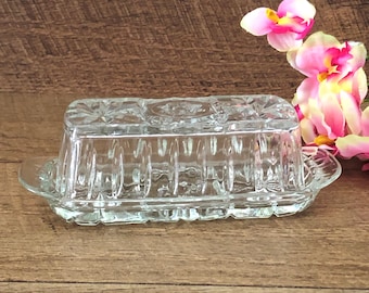 Op de afbeelding: Een glazen botervlootje met een decoratief patroon op de deksel en de zijkanten. De botervlootje heeft een rechthoekige vorm met een afgeronde rand en een klein handvat aan één kant.