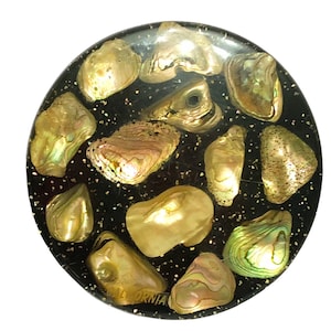 May include: Round, black coaster with a glittery surface and embedded iridescent seashells. The coaster has the word "California" written on the bottom.