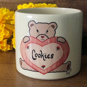 May include: White ceramic cookie jar with a red heart design. The heart has the word "Cookies" written on it. A brown teddy bear with a red heart is holding the heart.