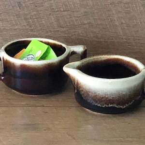 May include: A set of two brown and white ceramic creamer and sugar bowls with a glazed finish. The sugar bowl has a handle on each side and the creamer has a single handle.