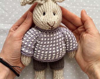 May include: A knitted beige bunny wearing a purple and white sweater and brown pants. The bunny is held in two hands.