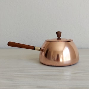 May include: A polished copper saucepan with a rounded body and a flat lid. The lid features a small, dark brown wooden knob handle. A long, straight, dark brown wooden handle is attached to the side of the copper pot with a metallic ferrule.