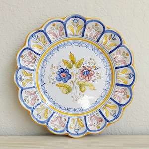May include: A round ceramic plate with a scalloped edge, featuring a white center painted with a blue flower and a pink flower surrounded by yellow leaves. The border has a yellow rim and blue and pink floral motifs on a white background.