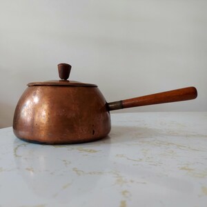 May include: A vintage copper saucepan with a wooden handle and lid. The pot has a rounded body and a small wooden knob on the lid. The handle is attached with a brass fitting. The copper has a warm, aged patina.