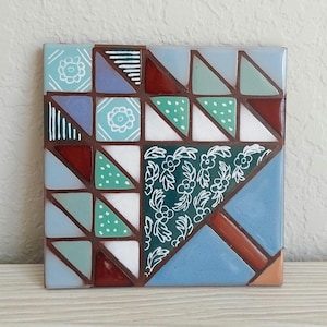 May include: A square ceramic tile featuring a geometric quilt-inspired pattern. The design consists of various triangles and squares in shades of blue, green, white, and dark red, accented with white line drawings of flowers, leaves, and dots.