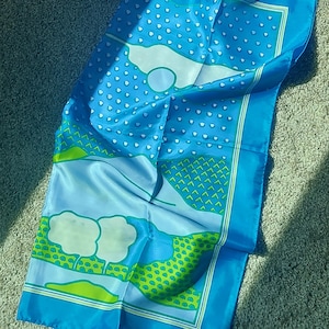 May include: A blue scarf with a white and green pattern. The scarf has a light blue background with white cloud-like shapes and small white dots. Green and white geometric shapes border the scarf.