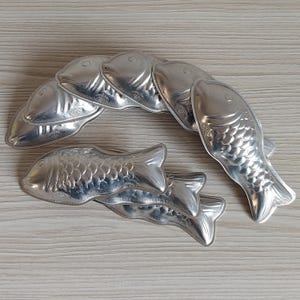 Könnte beinhalten: Eine Sammlung von silberfarbenen Metallformen in Fischform. Jede Form weist geprägte Schuppenmuster, einen definierten Kopf und eine Schwanzflosse auf. Die Formen sind in einer gebogenen Formation angeordnet und überlappen sich teilweise.