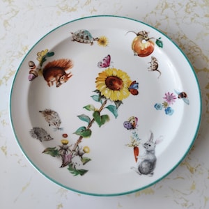 Puede incluir: Un plato de cerámica blanco con un borde verde presenta un diseño caprichoso de animales y flores. El plato tiene un girasol, una ardilla, un erizo, un conejo, mariposas y otros pequeños detalles.