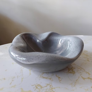 May include: A gray ceramic bowl with a unique, wavy shape. The bowl has a glossy finish with a speckled, textured rim. The interior is a darker shade of gray, creating a contrast with the lighter rim. The bowl is designed for decorative or functional use.