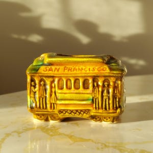 May include: A ceramic San Francisco cable car figurine in shades of yellow, green, and brown. The words "SAN FRANCISCO" are printed on the top. The figurine has detailed depictions of people and windows.