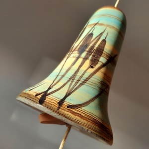 May include: A ceramic bell with a light blue, yellow, and brown striped design. The bell has a black painted design of tall grass.