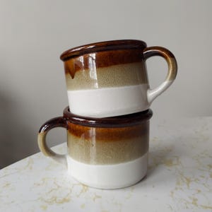 Puede incluir: Dos tazas de cerámica con asas, apiladas una encima de la otra. Las tazas son marrones, blancas y beige.