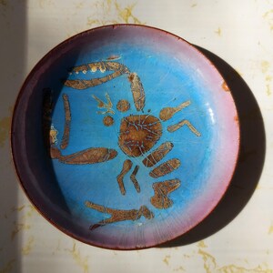Puede incluir: Plato decorativo redondo con centro azul y borde rosa. El plato presenta un diseño de cangrejo en tonos marrones y dorados. El diseño del cangrejo está en el centro del plato.