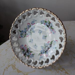 Puede incluir: Un plato de porcelana blanca con borde dorado y un delicado diseño calado. El plato está decorado con un patrón floral de nomeolvides azules y rosas.