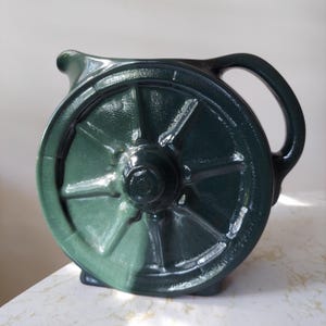 Puede incluir: Jarra de cerámica verde oscuro con diseño de rueda de carro. La jarra tiene un pico y un asa redondeada. El diseño de la rueda está en la parte delantera y trasera. Está colocada sobre una superficie blanca.