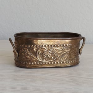 May include: An oval brass planter featuring a decorative embossed floral pattern along the exterior sides, a beaded border detail, and two small metal handles attached to each side. The metal surface has a warm, aged, and tarnished patina.