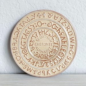 May include: A round, tan ceramic plate featuring the Cherokee alphabet embossed in concentric circles. The center of the plate displays the text "SEQUOYAH CHEROKEE ALPHABET" in clear, raised lettering, surrounded by the characters of the syllabary.