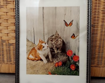 May include: Framed print featuring two kittens, one orange and white, the other gray, with butterflies. The kittens are near red flowers and green grass. The frame is silver with a decorative edge.