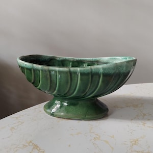 May include: A green ceramic planter with a pedestal base. The planter has a wide, oval shape with a fluted design on the exterior. The glossy finish gives it a vintage look.