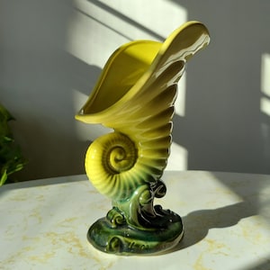 May include: A yellow and green ceramic seashell vase. The vase is shaped like a conch shell with a wide opening at the top. The base of the vase is decorated with green waves.