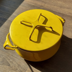 Puede incluir: Una olla holandesa de hierro fundido esmaltado de color amarillo brillante con una tapa a juego. La tapa presenta un asa distintiva en forma de cruz. La olla tiene dos asas curvas a cada lado. La olla está sobre una superficie de madera.