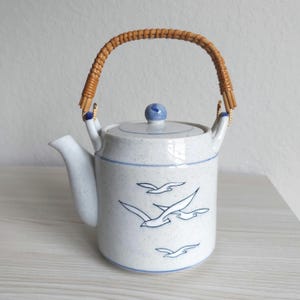 May include: A ceramic teapot with a speckled white finish and blue line accents. The body features a blue graphic illustration of flying seagulls. It includes a matching lid with a blue knob and a curved handle wrapped in natural brown rattan.