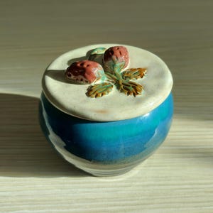 May include: A small round ceramic jar with a glossy blue and teal gradient glaze. The lid is off-white with a speckled texture and features a sculpted, hand-painted relief of two red strawberries with green leaves centered on the top surface.