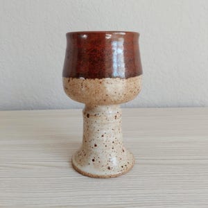 May include: A ceramic goblet featuring a two-tone design with a dark reddish-brown glazed upper bowl and a speckled cream-colored lower stem and base. The surface has a textured, speckled appearance throughout the lower section of the vessel.