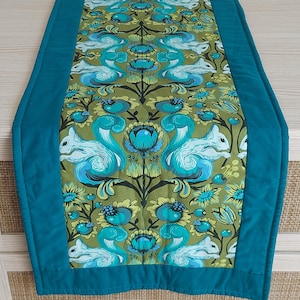 May include: A rectangular table runner featuring a central panel with a repeating pattern of light blue squirrels and teal floral motifs on an olive green background, framed by solid teal fabric borders with visible quilted stitching lines.