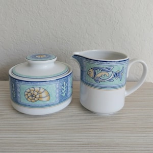 May include: A white ceramic sugar bowl with a lid and a creamer. Both pieces feature a blue and seafoam green border with seashell and fish designs. The sugar bowl is round, and the creamer has a handle and spout.