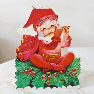 May include: Vintage Christmas ornament depicting a seated Santa Claus in a red suit and hat, playing a flute. The ornament has a white beard, glasses, and a gold belt. It is sitting on green holly with red berries. A gold string is attached for hanging.