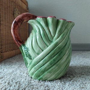 Puede incluir: Una jarra de cerámica con un diseño de hojas verdes. La jarra tiene un asa marrón y un borde con volantes con detalles en rojo. El cuerpo de la jarra es de color verde claro con sombras verdes más oscuras, lo que le da una apariencia texturizada.