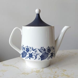 May include: A white ceramic teapot featuring a dark blue floral pattern around the base. The teapot has a curved handle, a tapered spout, and a removable dark blue lid topped with a small white spherical knob, creating a clean, functional design.