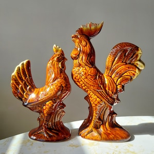 Puede incluir: Dos figuritas de gallo de cerámica, una más grande que la otra. Ambas son marrones con un acabado brillante y detalles elaborados. El gallo más grande tiene las alas extendidas, mientras que el gallo más pequeño está de pie con las alas plegadas.