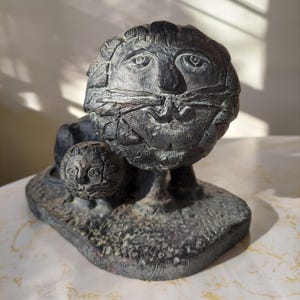 May include: A dark bronze sculpture of a lion and cub. The lion has a detailed face with a prominent moustache and a textured mane. The cub is smaller and positioned in front. The sculpture sits on a textured base.