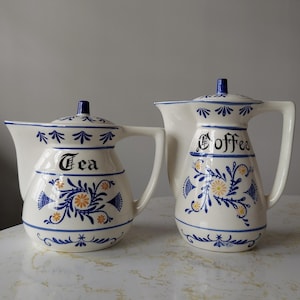 Puede incluir: Un juego de dos teteras y cafeteras de cerámica vintage con diseños florales azules y blancos. Las teteras tienen tapas y están etiquetadas como "Té" y "Café".