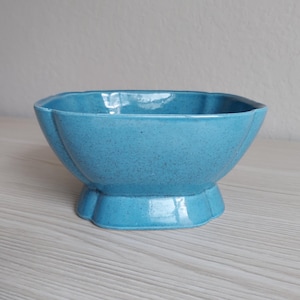 May include: A turquoise ceramic bowl with a speckled finish and a scalloped edge design. The bowl features a wide, flared opening that tapers down to a sturdy, pedestal-style base, creating a distinct silhouette with vertical ridges.