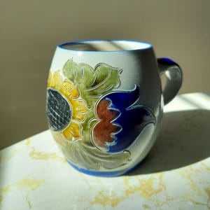 May include: A ceramic mug with a sunflower design. The mug is off-white with a blue rim and handle. The sunflower has yellow, green, brown, and blue petals. The mug is approximately 4 inches tall.