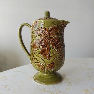 Può includere: Un'anfora in ceramica verde con un motivo floreale marrone. L'anfora ha un coperchio e una base a piedistallo.