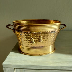 May include: A polished gold-tone metal bucket with a hammered texture on the lower body and horizontal ridges on the upper rim. The vessel features two side handles and a decorative twisted rope accent around the middle with two small tassels.