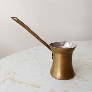 Puede incluir: Una cafetera turca de color latón con un mango largo. La cafetera tiene una base ancha que se estrecha hacia adentro y luego se ensancha hacia afuera hasta una parte superior más ancha. El mango está unido al costado de la cafetera.