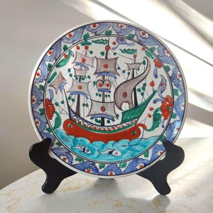 Peut inclure: Assiette décorative en céramique avec une illustration d'un voilier. Le fond est blanc, orné d'une bordure florale bleue. Le navire présente des voiles marron, une coque verte et des détails rouges, pour un thème marin.