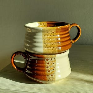 May include: Two stacked ceramic mugs featuring a two-tone design with a glossy cream-colored side and a rich brown side. The mugs have a horizontal ribbed texture and a speckled transition zone where the two colors meet, each with a curved handle.