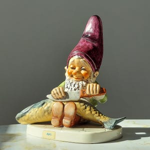 May include: A ceramic figurine of a gnome wearing a purple hat, holding a knife and sitting on a fish. The base of the figurine is white and has the word "FIPS" written on it.