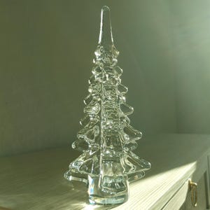 Puede incluir: Una escultura de vidrio transparente con forma de árbol de hoja perenne estilizado. El objeto presenta una punta lisa y afilada, con ramas redondeadas y escalonadas que se extienden desde una columna central vertical sobre una base circular.