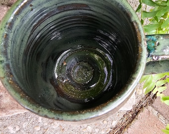 Op de afbeelding: Een groene keramische mok met een bruine glazuur en een gespikkelde afwerking. De mok heeft een brede, ronde opening en een handvat.