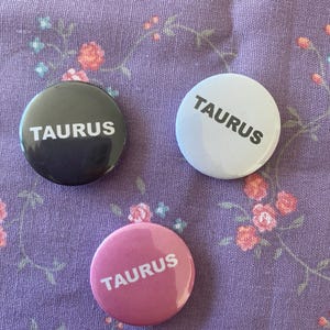 Puede incluir: Tres botones redondos con la palabra "TAURUS" impresa en blanco en cada uno. Los botones son negro, blanco y rosa.