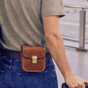 Può includere: Una borsa da cintura in pelle marrone con chiusura color oro, indossata su una cintura in pelle marrone sopra jeans blu. Uno zaino nero è visibile a sinistra. La persona tiene in mano il manico di una valigia.